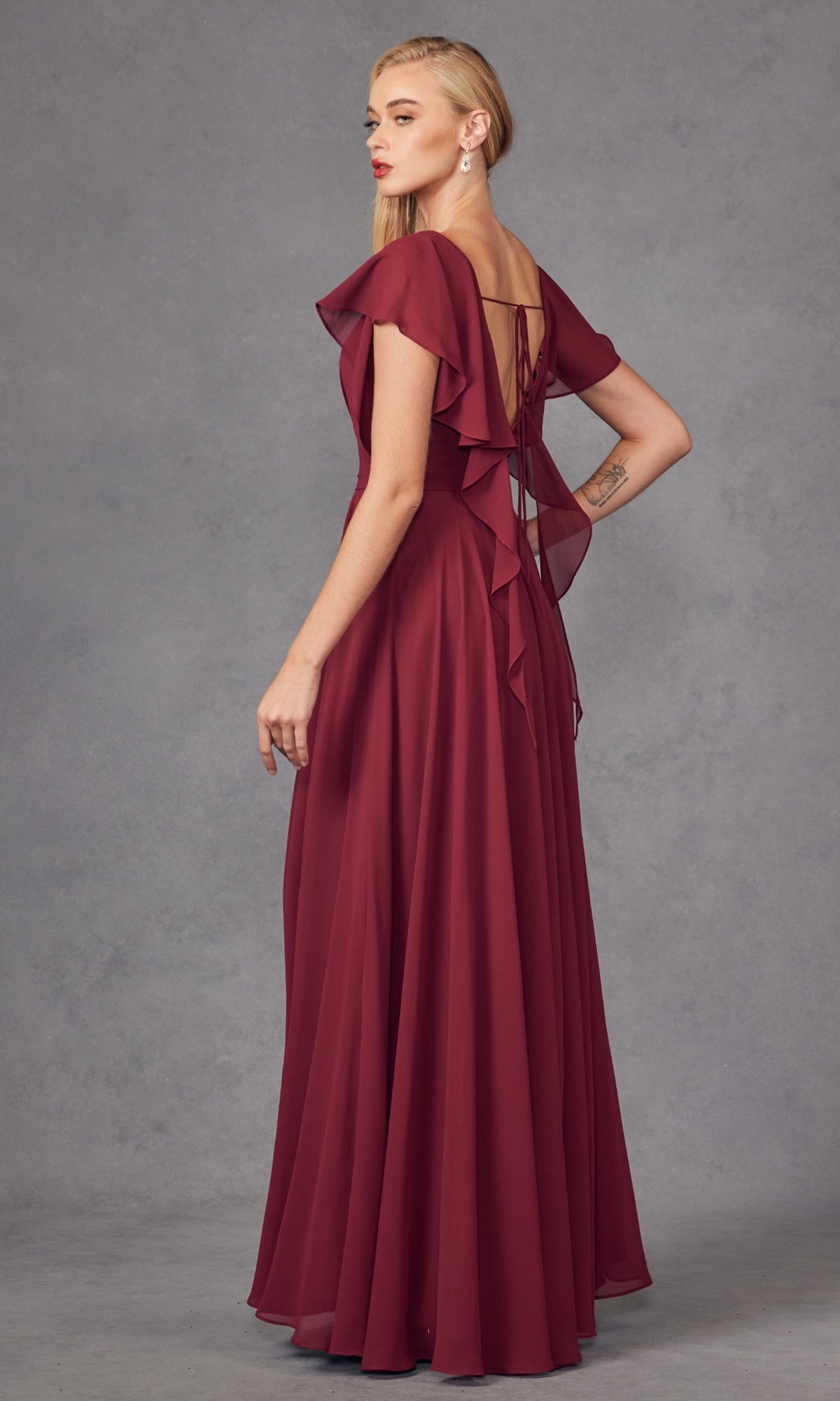 A woman in the Flutter-Sleeve Long Formal Evening Gown 261—a floor-length burgundy A-line dress with ruffle sleeves, lace-up back, and a flowing skirt—stands against a gray background.